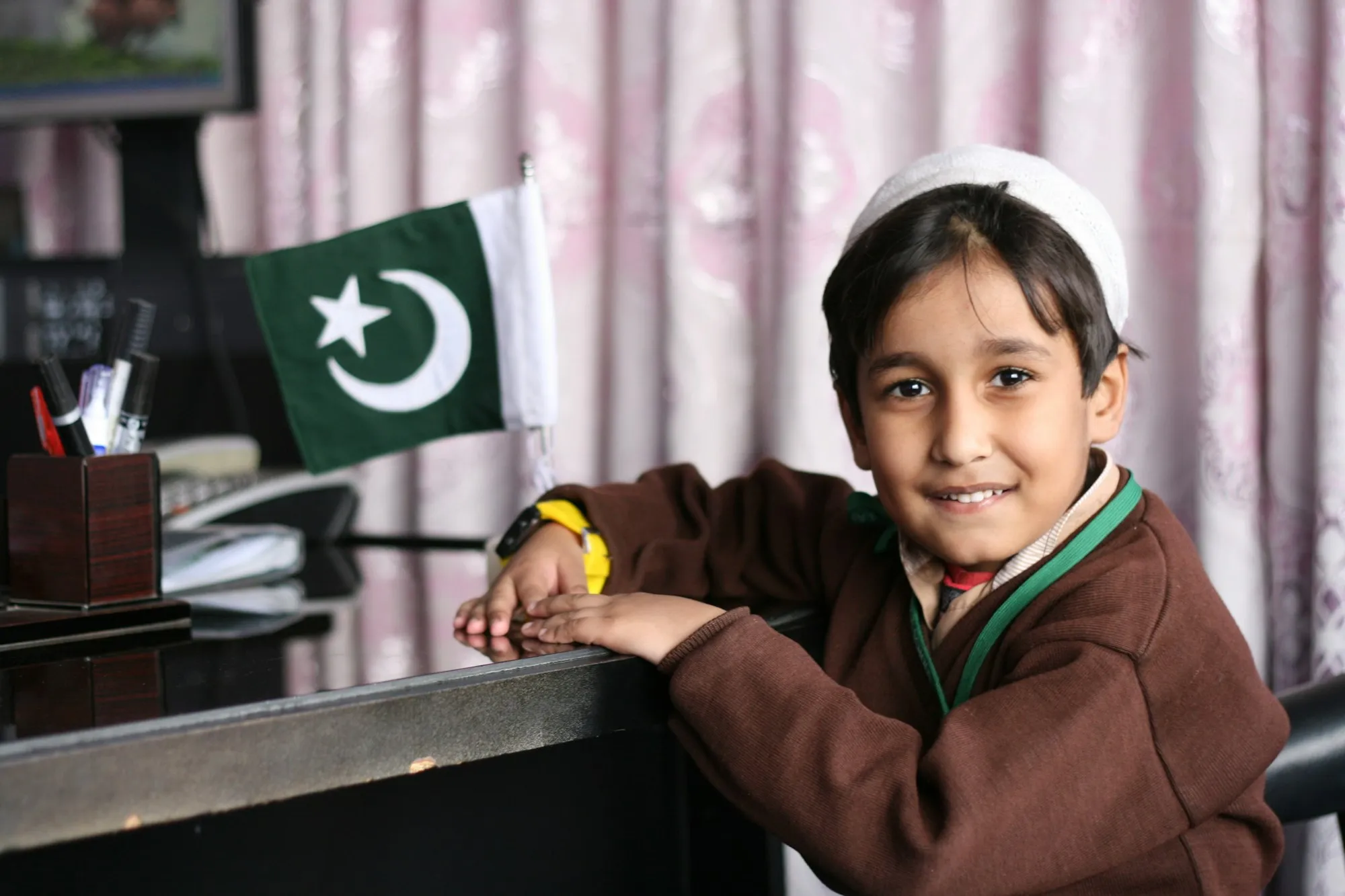 A young student in a brown school jumper at a desk with a small Pakistani flag